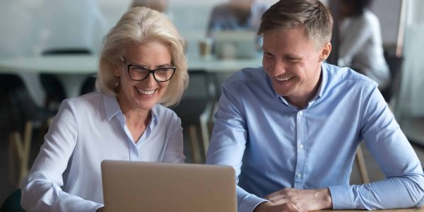 Joyful associates sitting coworking area using pc resting watching online funny videos on notebook. Experienced aged woman helping new worker understand corporate program people enjoy working together